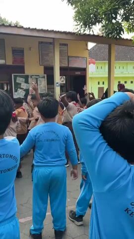 A group of young children are gathered outdoors, seemingly in a schoolyard. Many of them are wearing blue shirts with "SDN 2 TIRTOMOYO" printed on the back, and some are wearing brown uniforms, possibly scout attire. The children are facing a building with yellow walls and a covered walkway. A screen is visible on the building's exterior, displaying an image of several people performing some kind of synchronized movement, likely a dance or exercise routine. Many of the children have their arms raised, mirroring the poses on the screen. The environment appears to be warm, with trees visible in the background. The overall scene suggests an organized activity, possibly physical education or a school event, taking place outdoors.