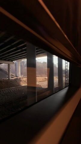 A view from inside a train through a window shows outside scenery. The train passes under a bridge. Train tracks and vegetation appear.