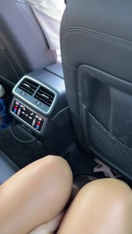 The video shows the interior of a car from the perspective of a passenger in the back seat. The focus is on the back of the front seats, specifically the air vents and climate control panel. There are two pairs of legs visible in the foreground, suggesting there are at least two people in the back seat. One person's bare foot is seen interacting with the climate control panel. The person's foot is placed on the panel, and their toes are seen moving and touching the controls. The climate control panel displays digital readouts for temperature and fan speed, with illuminated buttons. The environment appears to be a sunny day, as indicated by the lighting. The overall scene suggests a casual car ride where someone is playfully or deliberately adjusting the car's climate settings with their foot.