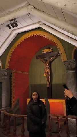 The video shows the interior of a church-like space that appears to be carved from rock. The architecture is unique, with a white, angled ceiling. In the first shot, a woman in a black coat stands in front of a colorful altar with a crucifix. The second shot pans across a group of people walking through the space. The third shot features a large, ornate chandelier hanging from the ceiling. Finally, the video focuses on a statue of a woman holding a child, positioned against a rough rock wall.