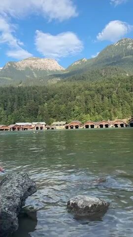 A body of water with rocks in the foreground. In the middle ground, a line of wooden boat houses with arched entrances is visible along the shore. Behind the boat houses, a dense green forest covers the lower slopes of mountains. The sky above is blue with scattered white clouds. The camera is positioned low, near the water's surface, giving the impression of being close to the water.
