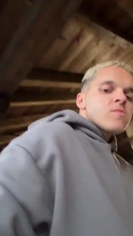 The video shows a young man with bleached blonde hair wearing a gray hoodie. He is in an outdoor setting with wooden tables and benches, suggestive of a restaurant or bar. In the background, there are two refrigerators, one with a Pepsi logo and the other a red vending machine. He appears to be speaking or singing to the camera. The lighting suggests it is either evening or an indoor/covered area.