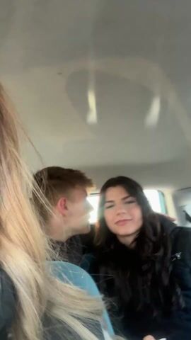 A blurry video shows two teenagers posing in the back of a car. The young man on the left has short brown hair and is wearing a black jacket. The young woman on the right has long, wavy dark hair and is also wearing a black jacket. She has white nail polish and long nails. The sun is shining behind them. The young man makes a silly face, sticking out his tongue and holding up his fingers in a peace sign. The young woman makes a kissing face. The seats are blue. A portion of a blonde head is visible in the foreground.