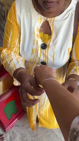 A woman in a yellow and white patterned dress is tying a thin white string around another person’s wrist. The woman has dark skin, gold jewelry, and a silver wristwatch. The person receiving the string has fair skin. A small red table is visible in the lower left corner of the frame. The scene appears to be taking place outdoors.