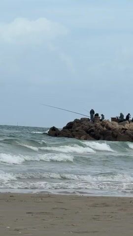 The video shows a group of people fishing from a rocky breakwater on a beach. The waves are crashing against the rocks, and the sky is overcast. There is a sandy beach in the foreground. Some fishing rods are visible. A tall lamppost is next to the rocks. A sailboat is visible in the distance.