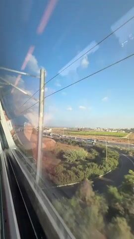 This is a video taken from inside a moving train, looking out the window at a landscape. The scene is dominated by the blue sky with scattered white clouds and several overhead power lines for the train. The window itself shows reflections, including what appears to be a person's arm and shoulder on the left side, and ceiling lights.

The view outside the window reveals a semi-rural setting. A highway with cars is visible in the middle ground, curving through an area with lush green trees and bushes. In the distance, there are fields and a city skyline, suggesting the train is passing through or near an urban area. The location is identified as Kfar Daniel, Israel.

The lighting suggests it is daytime, likely morning or afternoon, with bright sunlight. The weather appears clear and pleasant. The motion of the train causes a slight blur in the foliage closest to the window, indicating movement and speed. There are no visible text elements in the frame. The overall impression is one of travel and observing the passing scenery from a modern train.
