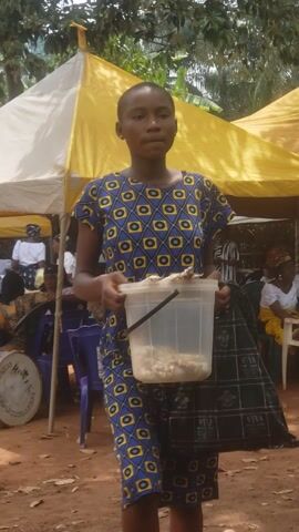 The video features a young person, likely a girl or young woman, standing outdoors in Umundugba, Nigeria, under a yellow fabric canopy. She has short, dark hair and is wearing a distinctive short-sleeved dress with a blue, yellow, and black geometric pattern. Throughout the video, she holds a clear plastic container with a black handle, which appears to be filled with a light-colored, granular substance, possibly a food item like popcorn or grains.

She looks directly at the camera at various points, maintaining a neutral expression. In the background, other people are visible, some seated on blue plastic chairs, suggesting a gathering or event. The ground is reddish-brown dirt, and green foliage indicates an outdoor setting. A white drum with the word "ROYAL" partially visible is on the ground in the background. The scene is brightly lit, indicating daytime. The activity primarily involves the individual holding and presenting the container while being filmed.