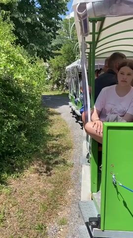 A train with open-air carriages moves along a path next to lush green foliage. People are seated in the carriages, facing forward. A young woman in a pink shirt is visible in the foreground. In the background, a mountain range is partially obscured by trees. The sun is shining, casting shadows on the path.