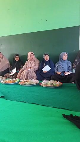 Five women are sitting on a green carpet, facing a wall that is green on top and dark green on the bottom. They are all wearing hijabs. The woman on the far left is wearing a light brown hijab and a patterned dress. The second woman from the left is wearing a black hijab and a patterned dress. The third woman from the left is wearing a light pink hijab and a patterned dress. The fourth woman from the left is wearing a dark blue hijab and a patterned dress. The woman on the far right is wearing a light blue hijab and a patterned dress. In front of the women are several platters of food. The food appears to be a variety of sweets and possibly some savory items. There are also some papers and books on the platters. The room appears to be well-lit.
