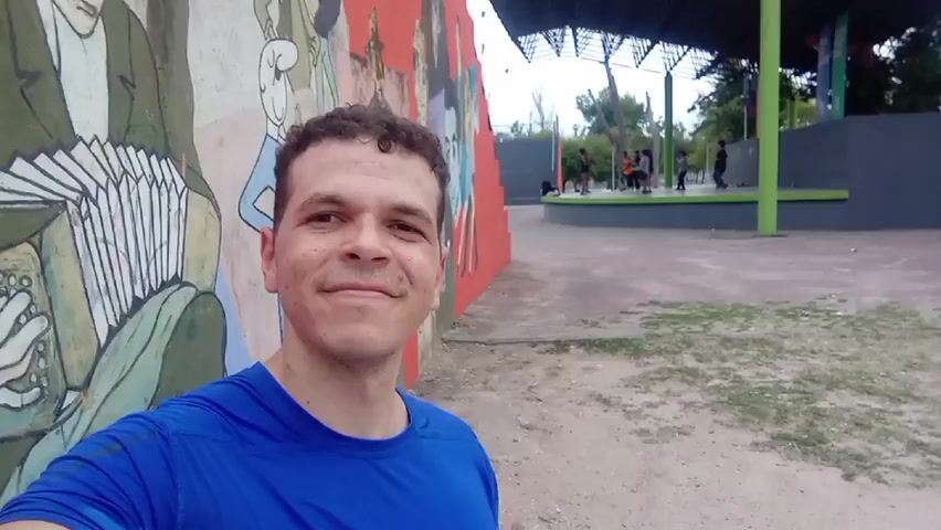 The video shows a man taking a selfie in front of a colorful mural, with a stage and several children visible in the background. The man is wearing a blue athletic shirt and has short, curly brown hair. He is smiling slightly at the camera. The mural is painted on a wall and features figures of people, including one playing an accordion. Behind the man and the mural is a small stage area with a dark gray platform and a dark gray roof supported by a green pole. There are several children playing on and around the stage. The ground is sandy with patches of grass.