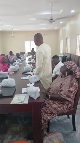 In a well-lit room with ceiling fans and fluorescent lights, a group of people are seated around a long, dark wooden table. A man in a light-colored traditional outfit stands at the head of the table, addressing the group and holding a microphone. The table is laden with white plastic bags, likely containing food or other items, and papers.

The attendees, some wearing headscarves and traditional attire, are attentive to the speaker. Their expressions suggest a formal gathering or a meeting. The room appears to be a conference room or a meeting hall, with windows providing natural light. The setting, with its simple furnishings and communal seating arrangement, points to an organized event.

The image captures a moment of discourse or presentation, with the speaker actively engaging the audience. The overall atmosphere seems serious and focused, indicative of an important discussion or distribution event. The presence of multiple white plastic bags on the table suggests a potential distribution of resources, perhaps food packages or supplies, within a community setting in Dadi Riba, Nigeria. The time of day is indeterminate, but the lighting suggests it is daytime.