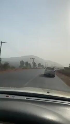 The video shows a car driving down a road, with a light-colored car ahead in the same lane.  The sky is bright, and there appears to be a hazy or smoky atmosphere.  Trees line the side of the road with power lines running along the roadside.  There is a mountain in the distance, obscured by the haze. The video is filmed from the perspective of the driver.