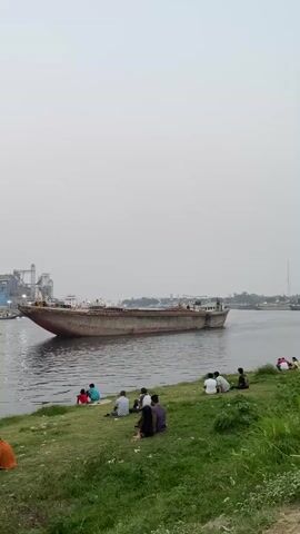 The video captures a tranquil scene by a waterway in Tarabo, Bangladesh. In the foreground, a lush green grassy bank slopes down to the water's edge, where a group of approximately ten to twelve individuals is seated. They are casually dressed and appear to be relaxing, observing the surroundings, or engaging in quiet conversation. One person on the far left has a bright orange bag placed next to them.

Dominating the midground is a large, long cargo vessel, likely a barge, slowly traversing the water. The boat appears somewhat weathered with visible rust, indicating its working nature. Faint, illegible text can be seen on its hull.

Across the water, in the background, an industrial complex is visible, featuring large buildings, silos, and machinery that resemble cranes or port infrastructure. The sky is a uniform light gray, suggesting an overcast or hazy day, likely during late afternoon or early evening given the soft, diffused light. The overall mood conveyed is one of calm and relaxation, with people enjoying the riverside ambiance and the passing boat.