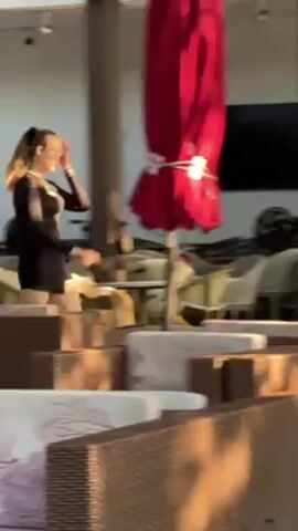 The video shows a woman in a black long-sleeved shirt and shorts walking through an outdoor seating area. She has her hair tied back in a ponytail. The area is filled with rattan furniture with white and patterned cushions. A large red umbrella stands nearby, and there's a display of purple orchids. An older man sits at one of the tables. The background features wooden planks and speakers. The scene is well-lit by natural sunlight.