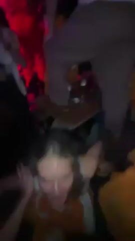 The video appears to be taken in a nightclub or bar setting, lit with red and pink lights. A woman with blonde hair is at the center of the frame, seemingly dancing or caught up in the crowd. There are other people around her, some with their arms raised, suggesting an energetic or celebratory atmosphere. The camera movement is erratic and close to the subject, contributing to the chaotic and dynamic feel of the scene. It is difficult to discern specific details due to the close proximity and low lighting.