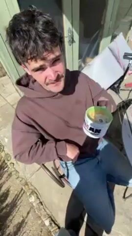 A young man sits on a chair outside, drinking from a mug. He wears a brown hooded sweatshirt and blue jeans. The mug is white with green accents and a design. He takes a sip, then gives a thumbs up to the camera. The setting appears to be a backyard or patio area, with stone paving and a green wooden shed in the background. The video is shot from a slightly elevated angle.