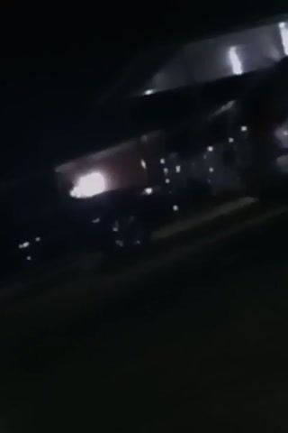 The video begins with a dark scene, showing a car with illuminated headlights under an overhang, followed by a brief pan to a multi-story building lit from within. The view then quickly shifts, becoming blurry, before focusing on a red scooter parked near a house. The scene is generally dark, with the primary source of light coming from interior lights of buildings.