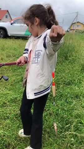 In a grassy outdoor setting, a young girl with curly brown hair is proudly holding up a small fish she has caught. She is wearing a white and blue letterman jacket over a red shirt, black pants, and white clogs. The fish is dangling from a fishing line, attached to a fishing rod held in her left hand. A red and orange fishing float is also visible on the line above the fish.

The girl appears happy and excited, with a smile on her face. She is looking towards the camera, showcasing her catch. The background shows a cloudy, overcast sky, suggesting it might be a cool or damp day. There are houses with red roofs visible in the distance, along with a white car with orange and green accents. The general scenery indicates a rural or suburban area, likely a field or meadow suitable for fishing. The location context places this scene in Bolshiye Polya, Russia.

The text visible on the girl's jacket includes "P" and "PLACES" written in a collegiate style.