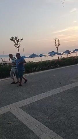 The video shows a walk-through of an outdoor area, possibly a promenade or park, near a body of water, likely a sea or lake, with the sky suggesting either sunrise or sunset. Several beach umbrellas are visible in the background. People are walking along a paved path. In the foreground, a child in a white shirt and a cap is walking. A man in a blue shirt and shorts is also walking. Further back, a person in a red shirt is seated on a bench. There are trees and palm trees lining the path, and trash bins are present. A dark statue, possibly of an elephant, is visible to the right. A section of a white wall with some decorative elements and a statue on top is also seen. The overall scene is casual and relaxed, with elements suggesting a public recreational space.