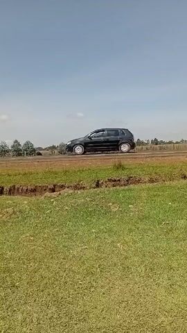 The video shows a view of a grassy field bordered by a road.  A black car drives along the road from left to right. The field is mostly green with some dry, brown patches. There is a small ditch or drop-off between the camera and the road. A line of trees can be seen in the distance on the other side of the road. As the camera pans, a long shadow appears on the grass, likely cast by the person holding the camera.  The sky is blue with some light clouds.