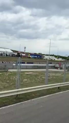 The video shows a race track on an overcast day. A grey road with a white line runs along the bottom of the frame, with a metal guardrail and chain-link fence between the road and the track. Beyond the fence is the track itself, lined with grey barriers featuring "LOUIS VUITTON" signage. In the background, there are white tents, a tall tower with red and white stripes, a blue truck with "MICHELIN" branding, and other trucks and vehicles. A racing vehicle speeds across the frame from right to left, creating a loud noise as it passes.