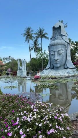 The video opens with a wide shot of a serene pond adorned with lily pads and blooming pink and white flowers in the foreground. Dominating the scene is a colossal stone statue of a face, likely representing a deity, with a serene expression. To the left of the face, a large stone hand with fingers extended upwards emerges from the water. A person is visible in the pond, kneeling near the large stone hand. In the background, lush green trees and palm trees sway under a bright blue sky with scattered white clouds. The camera pans to the right, revealing a pathway flanked by vibrant pink and white bougainvillea flowers. Further along the path, a white stone sculpture of a dragon is prominently displayed. The video captures a tranquil and spiritual atmosphere, showcasing impressive sculptures set within a beautiful natural landscape.