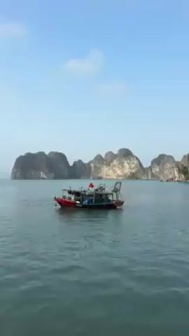 The video shows a red and black fishing boat floating on calm, greenish-blue water. The boat has a Vietnamese flag waving on it.  Behind the boat are several large, rocky islands with steep cliffs covered in vegetation. The sky is clear and pale blue with a few small clouds.