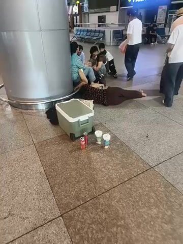 The media file shows a group of people at what appears to be an indoor transportation hub, possibly an airport or bus station in Thuận An, Vietnam. The lighting suggests it is daytime.

In the foreground, a woman in a patterned dress is lying on the floor next to a large, green and white cooler. Several cans of soda and cups with lids are placed in front of the cooler. Scattered around her are bags and clothing items.

To the left, a group of people are seated or leaning against a large cylindrical pillar. One woman in a light blue top is interacting with a child. Another person, possibly male, is leaning over the child.

In the background, two individuals are standing. One woman in a white t-shirt and black pants is holding a red plastic bag. Another person wearing a white t-shirt, dark pants, and a hat is carrying a guitar case and a dark garment over their shoulder. Other people can be seen in the distance sitting on benches in a waiting area.

The overall scene depicts people appearing to rest or wait, with some lying on the floor, suggesting a long period of waiting or potential hardship. There is no overt indication of distress, but the setting and the sleeping individuals might imply a need for rest or shelter.

Visible text on a sign in the background reads "PAM PHÔ VIET NAM" and "Coffee". Another sign has the ASCO logo.