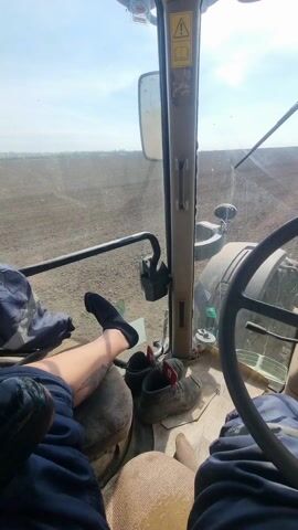 The video provides a first-person perspective from inside the cab of a tractor, looking out over a vast agricultural field. The scene is set in Dobrevo, Bulgaria, under a bright daytime sky with scattered clouds.

Inside the tractor cab, a person is visible with their legs casually propped up on the left side of the dashboard or seat, wearing black socks. A tattoo is visible on their left lower leg. A pair of dark shoes rests on the floor in front of them. The prominent steering wheel, adorned with the iconic John Deere deer logo, dominates the right side of the frame, indicating the tractor's brand. Various dashboard controls and levers are also visible. On the overhead panel, a yellow triangular warning sign with an exclamation mark is clearly seen.

Outside the windshield, the front part of the green tractor and its large tires are visible. The land stretching ahead is a wide, open field of dark brown, freshly tilled soil, suggesting preparations for planting or other agricultural activities. The field extends to the horizon under the bright sunlight. The tractor appears to be in motion, navigating across the expansive tilled land. The overall impression is one of routine farming operations taking place on a sunny day.