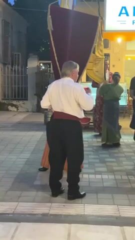 The video begins with a man in traditional attire, playing a flute. He is dressed in a white shirt, black pants, and a red sash. He is facing away from the camera and towards a group of people standing in front of a restaurant. The restaurant's sign, "ANDREAS RESTAURANT," is visible. A woman in a red and gold dress is standing to the left of the flutist.

The scene then shifts to show a group of women in traditional clothing, characterized by red and white striped dresses and white socks. They appear to be gathered and interacting. One woman in the center is holding a bottle of water. A woman in a black outfit is standing to the right, facing away from the camera. The background shows a bustling street scene with more people and lights from the restaurant. The pavement is made of interlocking rectangular stones. The time elapsed in the video is 5 seconds.