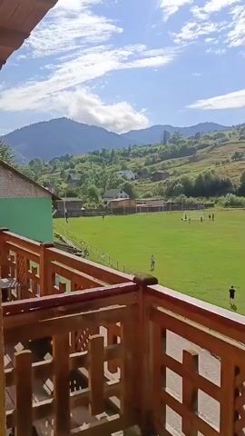 The video shows a daytime view from a wooden balcony overlooking a green sports field. Several people are visible on the field, some of whom appear to be playing soccer. In the background, there are rolling green hills and mountains under a partly cloudy blue sky. The balcony railing, made of dark wood with a lattice design, is in the foreground.