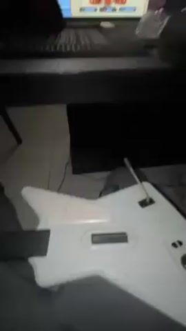 A first-person perspective video shows a person holding a white guitar controller and sitting in front of a television screen displaying a game with a cartoon-like interface. The room is dimly lit, and the camera pans upwards to reveal a table with various items, including a keyboard, cups, and other unidentified objects. The room appears to be a living area with a staircase visible in the background.