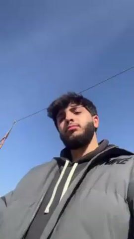 The video shows a young man outside on a sunny day. He is wearing a gray jacket with a Nike logo and a black hoodie. He makes a peace sign with his hand. The camera rotates to show several parked cars, a building made of cinder blocks, and a red water tank on the roof. The background is blurry, but there are trees and power lines visible. There is a man walking in the background. Construction or repair activity appears to be taking place on the building.