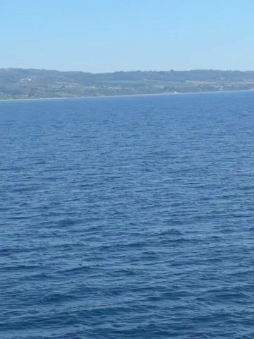 The video shows a calm blue sea with a distant coastline. The coastline is characterized by rolling hills covered in green vegetation, with some scattered buildings and houses visible. The sky above is clear and blue, suggesting a sunny day. The camera appears to be positioned on a boat or from a higher vantage point looking towards the shore. There are no people or specific objects clearly discernible on the water or the land due to the distance and resolution of the footage.