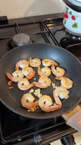 The video shows a close-up view of shrimp being pan-fried in a black skillet on a gas stovetop. The shrimp are arranged in a circular pattern within the skillet, and they appear to be a light pinkish-orange color, indicating they are cooking. Several pieces of what look like minced garlic are scattered among the shrimp. The skillet is placed on a black gas stove with four burners. In the background, a wooden countertop is visible, with various items including a white pitcher, a bottle of olive oil, a jar of pickles, a cutting board, and some plates with food. The overall environment appears to be a kitchen setting. The video has a duration of 4 seconds.