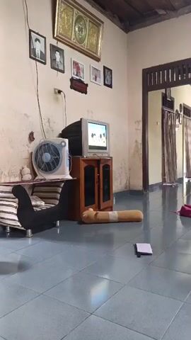 A low-angle, static shot captures a living room with a tiled floor. To the left, a white oscillating fan rests on a striped couch, partially obscured by a dark, slinky-like object. A small television set sits atop a wooden cabinet in the center of the frame. To the right of the cabinet, a long, tan cylindrical pillow lies on the floor. A small, white notepad is also on the floor in the foreground. Further back, a doorway reveals a hallway with curtains. Two children are visible in the doorway: a girl in a light-colored, patterned dress stands facing right, and a boy in a blue shirt sits on the striped couch. The wall behind them is painted a light color and shows signs of age. The ceiling is wooden.