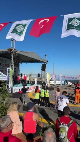 The video shows an outdoor event with a crowd of people gathered. There are flags of Turkey and other banners, suggesting a national or local event. Some individuals are wearing athletic attire, and there are signs indicating a "START" area and "AMONOS ULTRA TRAIL". People appear to be milling around, possibly preparing for a race or event. There are tents and a stage set up in the background. The sky is clear and blue, indicating fair weather. The overall scene suggests a start line or gathering point for a running or outdoor sports event.