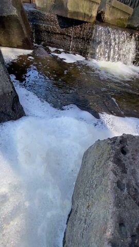A small waterfall cascades between concrete structures and large rocks, creating abundant white foam as the water rushes through the narrow space. The water is dark, and the surrounding area appears to be outdoors, likely a constructed water feature in a park or garden.