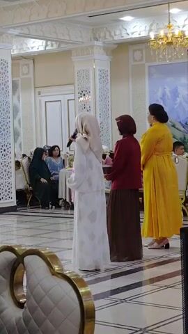The video shows an indoor event with people seated at tables and standing in an open area. The room appears to be a banquet hall, decorated with ornate columns, chandeliers, and wall art. In the foreground, a man is seated, looking towards his right. In the middle ground, several women are seated at tables, some of whom are wearing headscarves. A woman in a white outfit and headscarf is standing and speaking into a microphone. To her right, two other women are standing. In the background, more guests are seated at tables, and some individuals are standing and walking around. There are also decorative elements like floral arrangements and lights. The overall atmosphere seems to be that of a celebration or a formal gathering.