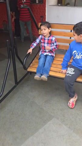 Here is a description of the video:

Two young boys are sitting on a wooden porch swing. The boy on the left wears a red and blue plaid shirt and blue jeans. The boy on the right wears a blue sweatshirt with "Chicago 1998" on the front and grey sweatpants with "Bugs Bunny" written on the leg. He also wears colorful sandals. The swing is made of dark metal and light wood. The background is a white wall with a red painted section. The older boy says something while the younger boy listens.