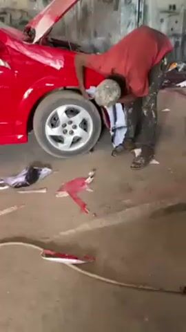 A man in a red shirt, camouflaged cap, and paint-splattered pants is crouched next to the front left wheel of a red car with its hood open. He is wiping the fender with a white cloth. Paint-covered cloths and parts litter the floor around him and the car. He is inside a garage or workshop.