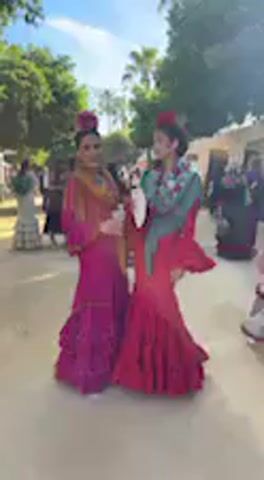 Two women in traditional flamenco dresses are the central subjects of the video, engaged in conversation and smiling. The woman on the left wears a vibrant magenta dress with ruffled layers and a multi-colored shawl in shades of orange and gold. Her hair is styled up with a flower ornament. She holds a small white object, possibly a fan or a drink container, and appears to be speaking. The woman on the right is dressed in a striking red flamenco gown with extensive ruffles and a green and blue patterned shawl, also adorned with a flower in her hair. She is looking at the other woman and smiling.

In the background, other people are visible, some also in similar elaborate dresses, suggesting a festive gathering. The setting is outdoors, under a clear, bright sky, indicative of daytime. Trees and what appear to be tents or temporary structures are scattered throughout the background, characteristic of a fairground or outdoor event. The overall atmosphere is lively, with background chatter and Spanish music (possibly flamenco or sevillanas) audible, fitting the context of Jerez de la Frontera, Spain. The women appear happy and relaxed, enjoying the event. No discernible text is visible in the video.
