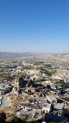 This is a vertical, wide-angle aerial shot of a desert landscape with peculiar rock formations. In the foreground, there are stone buildings with red tiled roofs. A white minaret rises from one of the buildings. The terrain further away is a mosaic of arid, rocky hills and valleys, dotted with more of the strange, conical rock formations. The sky is clear and blue. The scene appears to be a settlement in a geologically unique area.