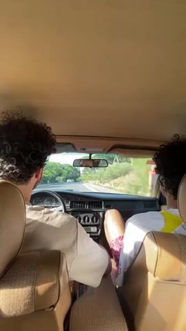 The video shows two people in a car, viewed from the back seat. The person in the driver's seat wears a white shirt and has curly dark hair. The passenger wears a white shirt with a yellow patch and has curly dark hair. The car appears to be an older model with a beige interior. The car is moving along a road lined with green trees and vegetation. In the distance, other vehicles can be seen on the road. The scene appears to be bright and sunny.