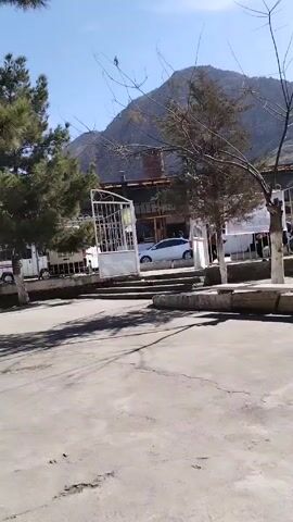 The video shows a view of what appears to be a town square or courtyard on a sunny day. In the background is a mountain range with a clear blue sky above. 
In the middle ground, there is a white metal gate with steps leading up to it. People can be seen walking through the gate. There are also cars and vans parked behind the gate. To the left of the gate, there is a green pine tree. To the right, a bare tree stands.
The foreground shows a concrete surface with cracks and shadows. 
The camera is static.