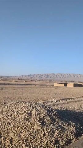 The camera travels along a paved road on a clear, sunny day. To the side of the road are piles of gravel and a flat, dry landscape with sparse vegetation. In the distance, a range of brown mountains extends across the horizon. There are a few simple, rectangular structures visible in the middle ground, made of earth-toned material. The sky is a bright, cloudless blue. There are no people visible in the video.