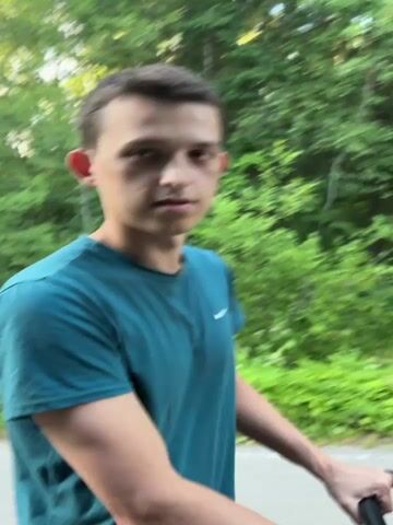A young man with short brown hair, wearing a teal t-shirt, is seen from the chest up, looking towards the camera. He appears to be outdoors in a natural setting, with lush green trees and foliage in the background. The lighting suggests it is daytime, likely afternoon, with soft, diffused light. The man's expression changes throughout the short clip, from a neutral gaze to a wide smile with visible teeth, and then to a more neutral, slightly pensive look. He is holding onto what appears to be the handlebars of a bicycle or stroller. The setting is identified as Shevlyagino, Russia. There is no visible text in the media.