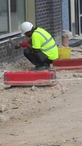 A person is working on a construction site. They are wearing a white hard hat, a neon yellow safety vest with reflective stripes, dark pants, and red gloves. The person is crouched down and appears to be using a tool, possibly a trowel, to apply material to a brick wall. The background shows a brick building, a window, a yellow bucket, and red construction barriers. The ground is covered in gravel and debris. The scene suggests it is daytime, and the weather appears to be fair. There is no visible text in the image.