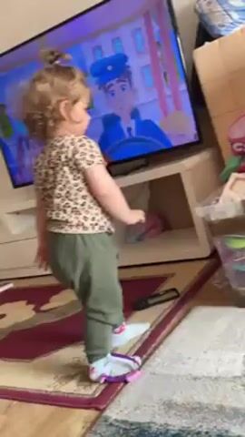 A young, light-skinned child with curly blonde hair in a high bun is standing on a burgundy and white rug in a living room, facing a large flat-screen television. She is wearing a beige and brown leopard print shirt and loose-fitting olive green pants with blue socks. The child is holding a rectangular white picture frame in her hands and tapping the frame on the rug. The TV is playing a cartoon featuring a blue uniformed character driving a vehicle. A white TV stand sits below the TV, with a DVD case on top. To the right of the child and TV stand, there's a cardboard box, possibly containing items, and a clear plastic storage container filled with various craft supplies.  A pair of purple scissors are lying on the rug in front of the child.