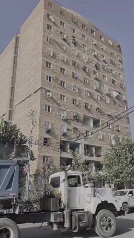 A tall, multi-story apartment building with visible damage, including missing walls and broken windows, stands under a clear sky. A large blue dump truck is parked in the foreground, partially obscuring the lower section of the building. Trees and foliage grow around the base of the building. Power lines are visible crisscrossing in front of the structure. In the distance, a white SUV and two individuals walking on a sidewalk are present. The overall scene suggests a damaged urban environment.